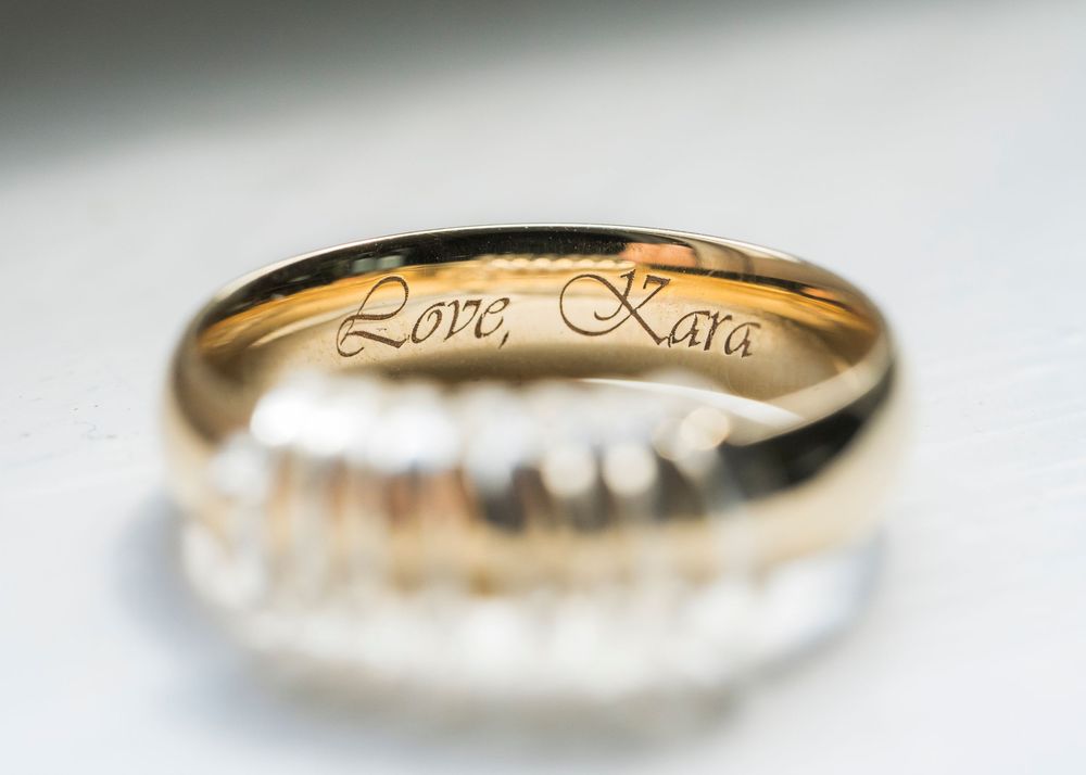 A gold wedding band with Love Kata engraved on the inside sits on a reflective surface creating a delicate shadow.