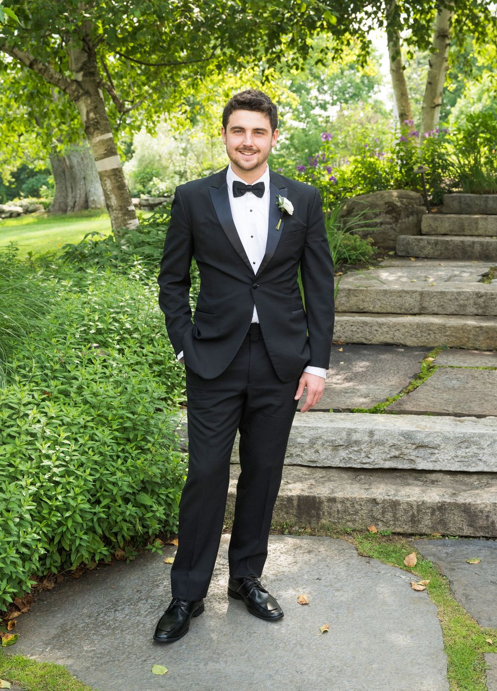 A dapper wedding party member in a black tuxedo and bow tie poses on stone steps in a lush garden setting.