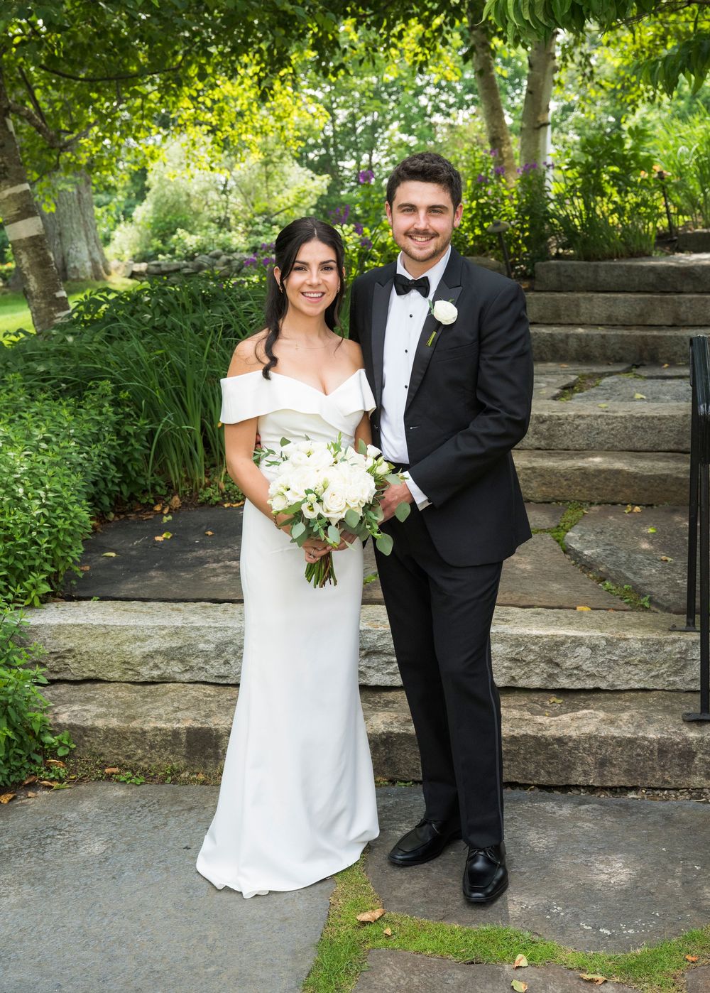 Wedding couple poses on stone steps in a lush garden setting with white bouquet and formal attire.