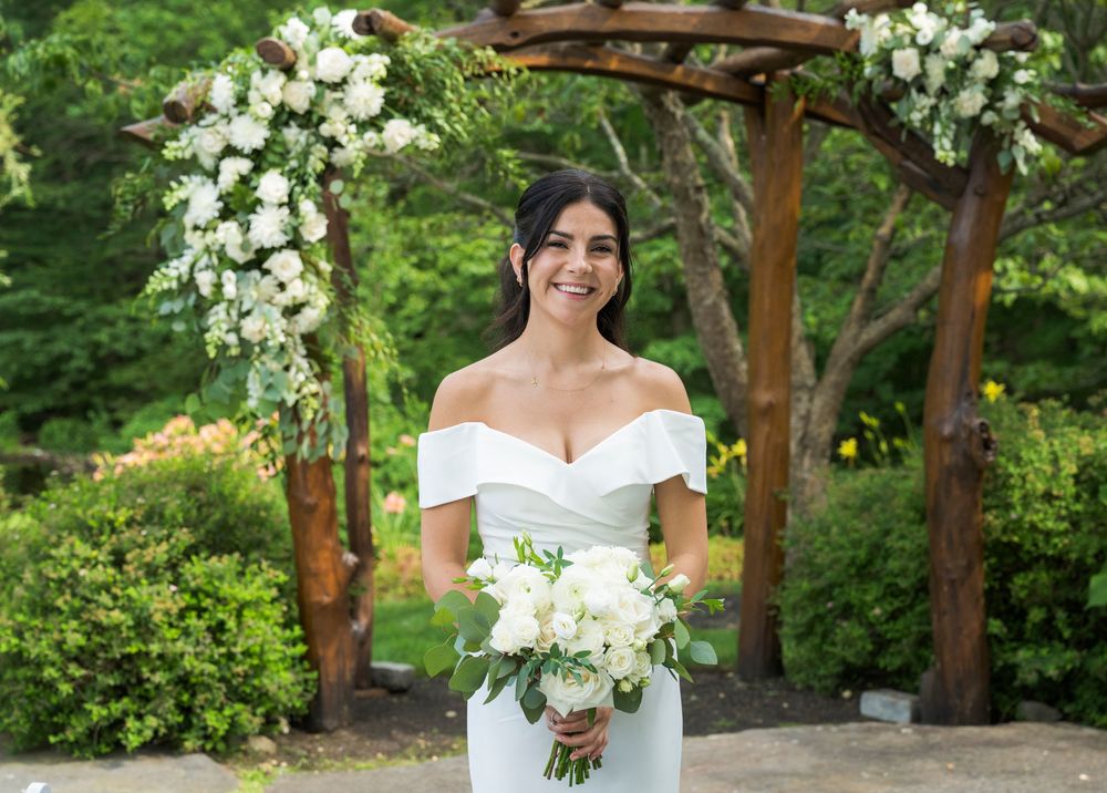 A bride poses under a rustic wooden arch adorned with cascading white flowers in a garden setting.