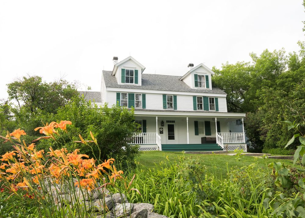 White colonial style house with wrap-around porch and orange daylilies blooming in foreground.