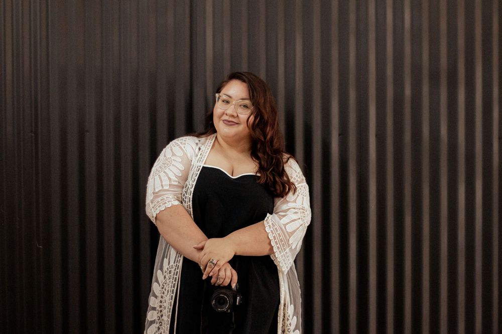 A plus-size woman wearing a black dress and sparkly silver cardigan poses confidently against a dark striped wall.