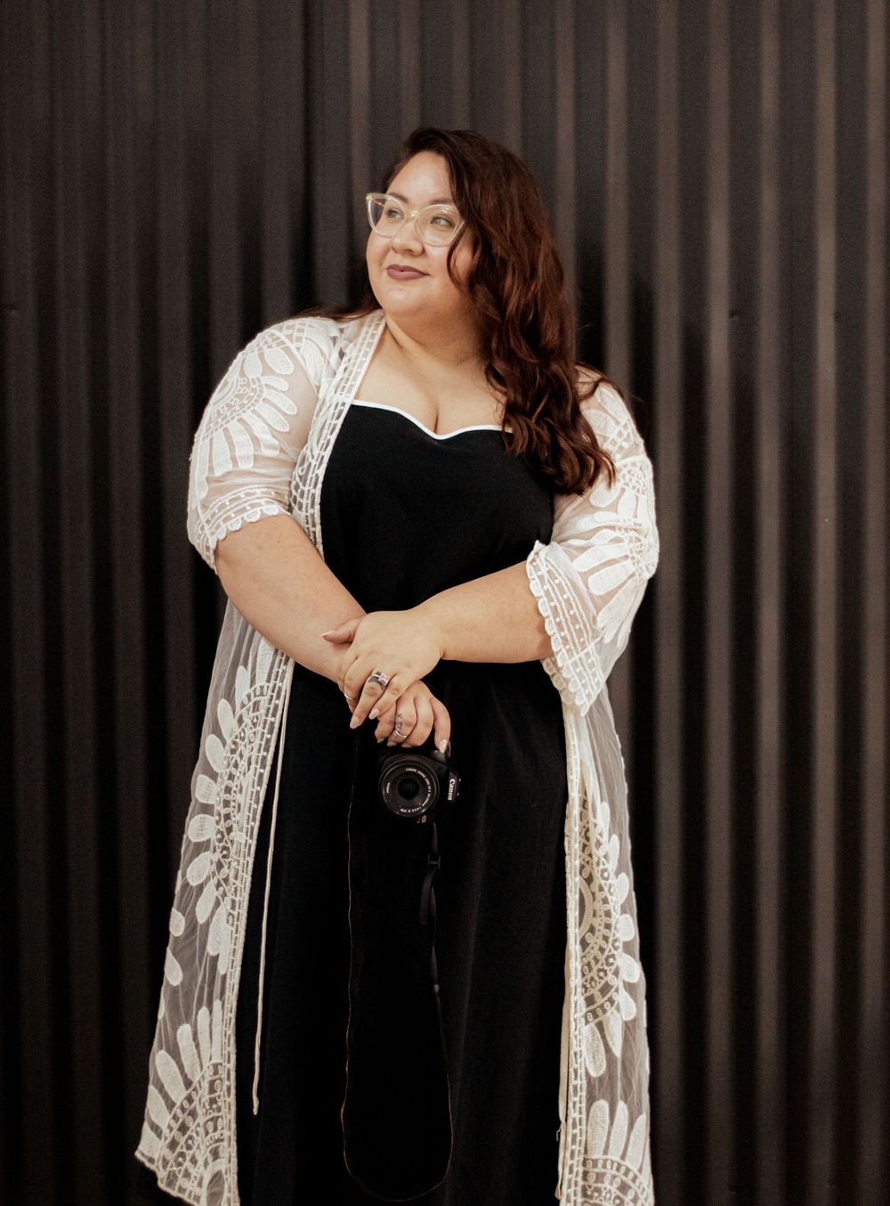 Curvy woman wearing black dress and lace overlay kimono poses confidently against dark striped wall while holding camera.