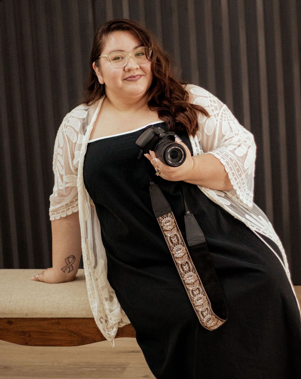 Photographer sits on wooden bench wearing black dress and lace overlay while holding professional camera.