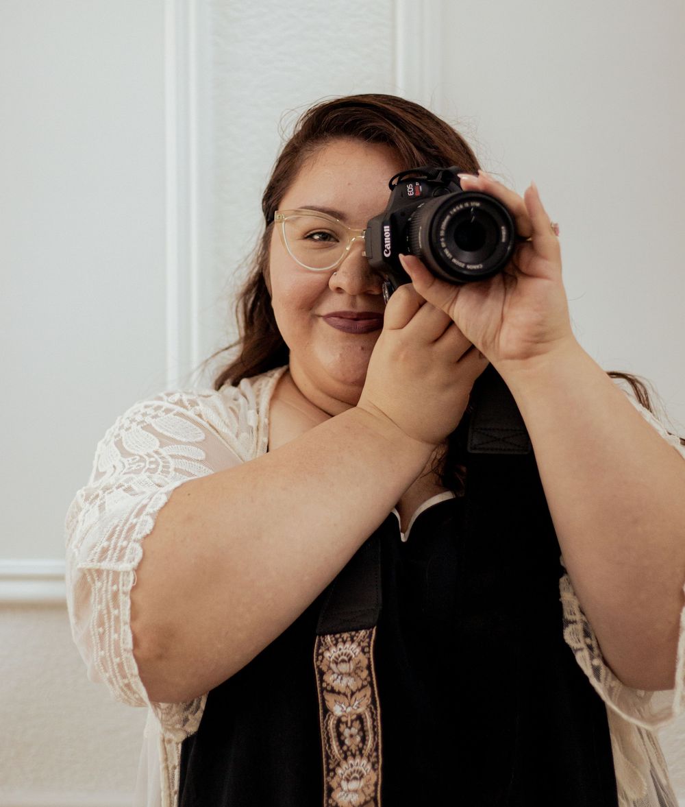 A photographer in a white lace top holds a black camera up to take photos in a light-filled studio space.