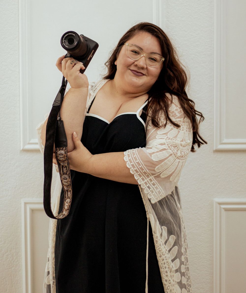 A photographer in a black dress and lace cardigan laughs joyfully while holding a professional camera.