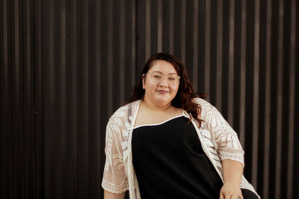 Person in a white lace top and black dress poses in front of dark striped wall with dramatic lighting.
