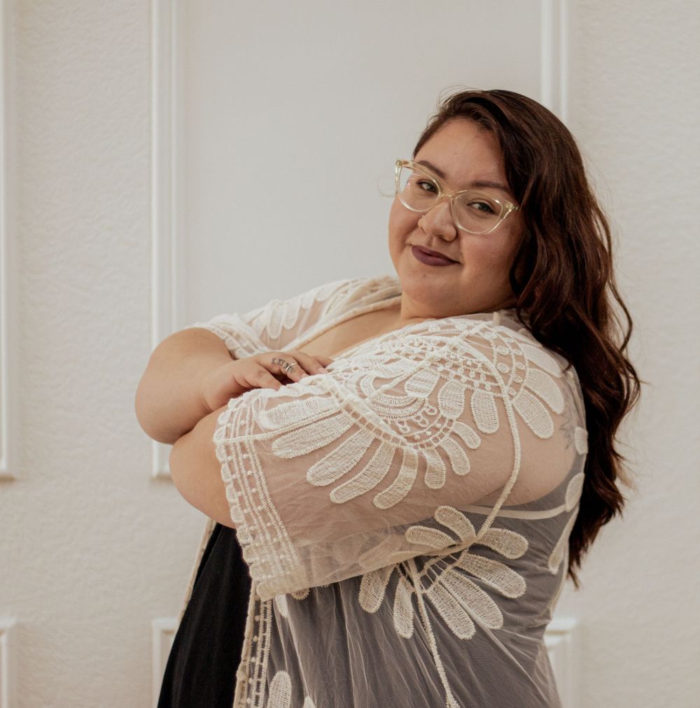 A side-posed portrait in a lace-detailed white dress against a light wall, showing a warm genuine smile.