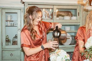 Person in salmon-colored satin robe pouring coffee into a cup in rustic kitchen with vintage cabinet.