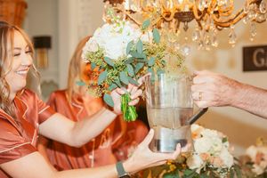Bridesmaids arranging floral bouquets under crystal chandelier at wedding preparation.
