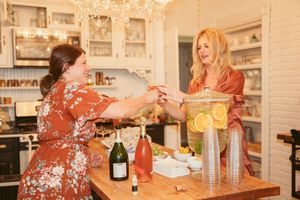 Two women in floral dresses preparing drinks and snacks in an elegant white kitchen.