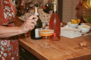 Hand holding a glass of orange cocktail on wooden counter with champagne bottles and snacks in background.
