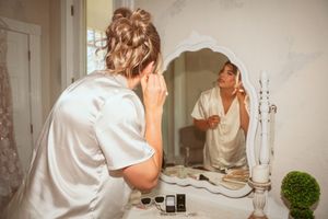 Person in white robe getting ready at bathroom mirror with cosmetics and toiletries.