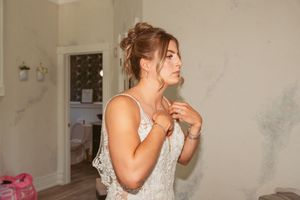 Woman in a white wedding dress adjusts her jewelry while getting ready in a dressing room.