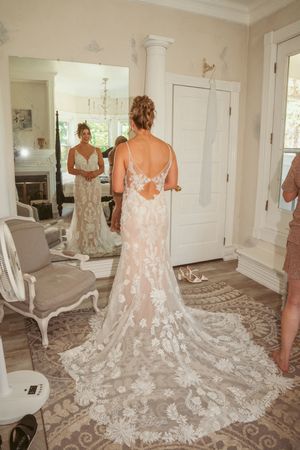 A wedding dress with intricate lace details and a dramatic cathedral train is displayed in an elegant white room setting.