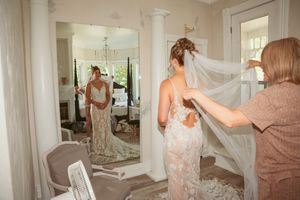 A person wearing a lace wedding dress gets help adjusting their long veil while looking in a mirror.