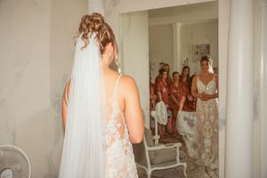 Back view of bride in lace dress and veil with bridesmaids reflected in mirror during wedding preparations.