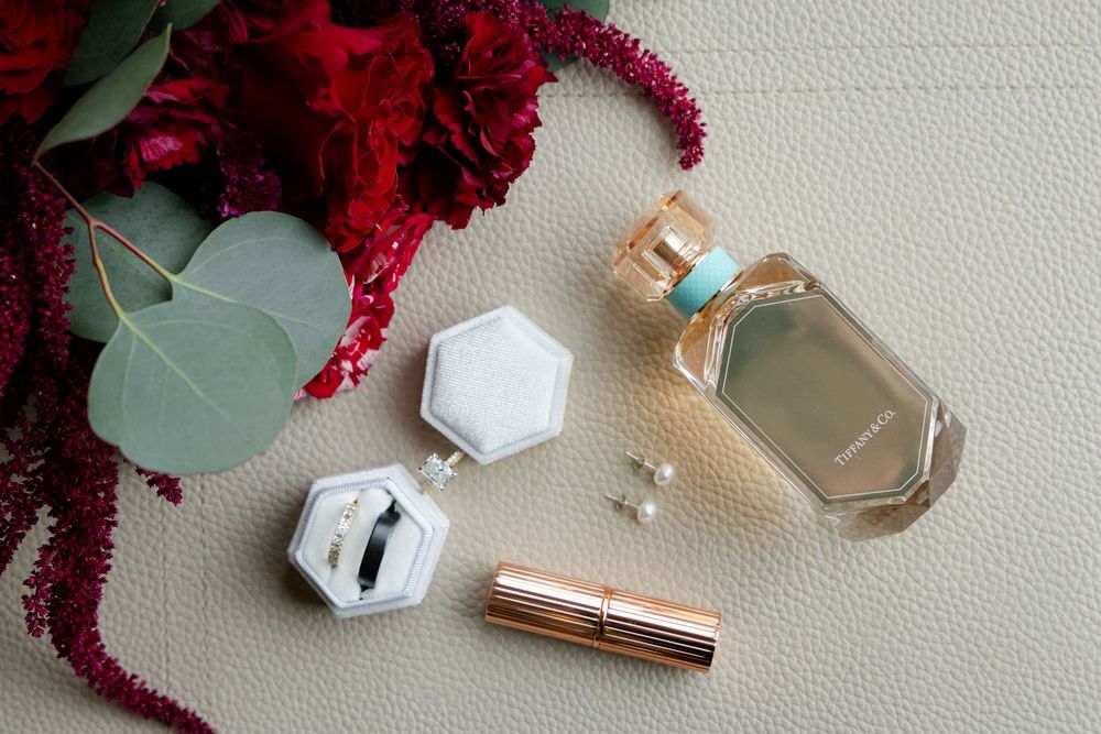 Wedding detail flatlay with perfume bottles, jewelry, and deep red floral accents