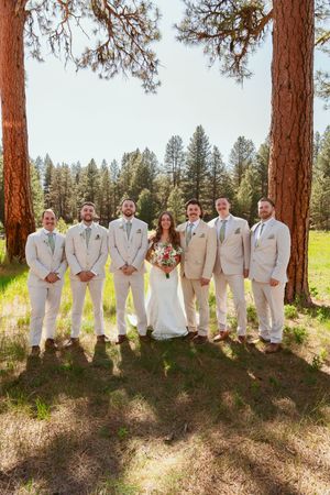 Wedding party in tan suits and white dress lined up along pine trees in natural outdoor setting.