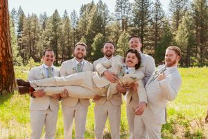 Groomsmen in beige suits pose playfully in a sunny meadow with pine trees in the background.