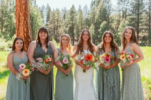 Wedding party in sage green dresses holding colorful bouquets pose in forest meadow.