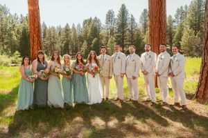 Wedding party members pose in long gowns and suits among tall rustic pine trees in an outdoor forest setting.