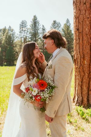 Wedding couple embraces between rustic tree trunks with colorful bouquet and natural lighting.