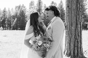 A black and white wedding photograph captured among tall pine trees in a serene forest setting.