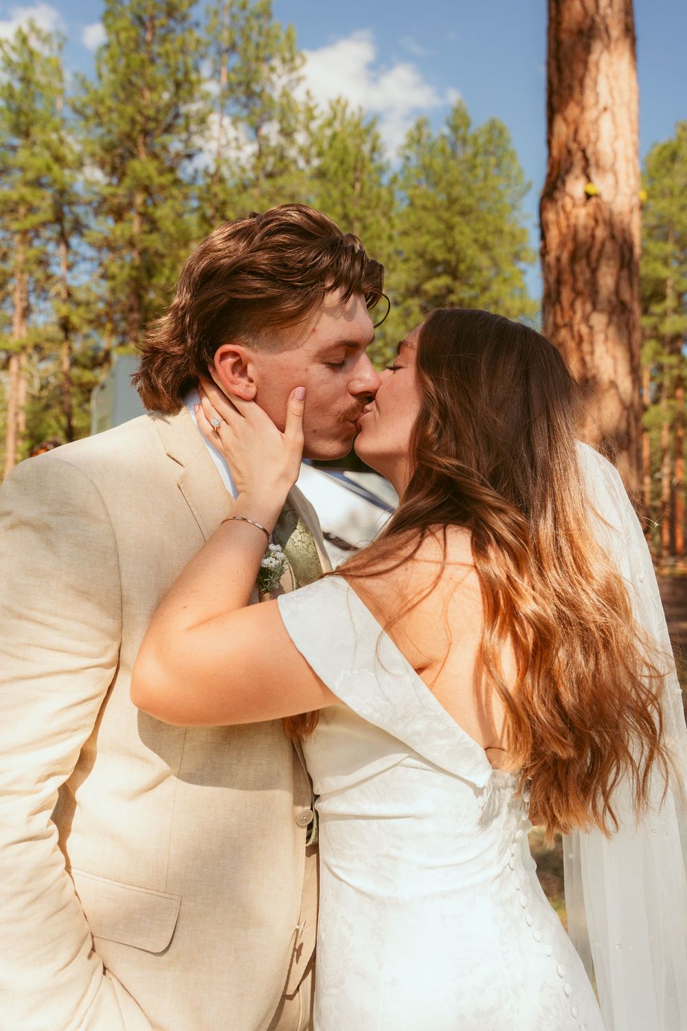A romantic wedding kiss between newlyweds in white and beige attire against a backdrop of pine trees.
