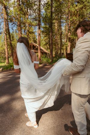 A bride in flowing white dress and sneakers walks down a wooded path as her dress train flows in the breeze.