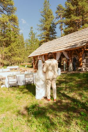 A rustic outdoor wedding reception setup with wooden cabin and dining tables surrounded by tall pine trees.