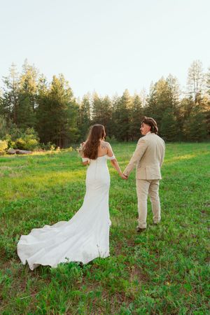 Couple in matching cream outfits dance and twirl playfully in a sunlit meadow with pine trees in the background.
