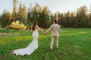 Two people hold hands while walking through a sunlit meadow surrounded by tall pine trees at golden hour.