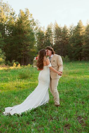 Couple dances and twirls in a grassy field with pine trees in the background at sunset.
