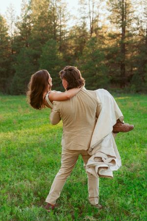 A romantic couple sharing a tender moment in a grassy meadow surrounded by tall pine trees at sunset.