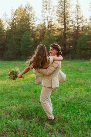 Two people in light colored attire dance together on a grassy field at sunset with evergreens in the distance.
