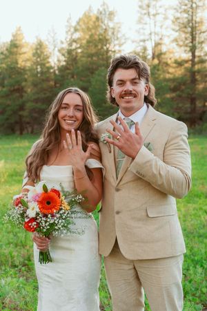 A happy couple shows off their wedding rings in a scenic forest setting with vibrant bouquet.