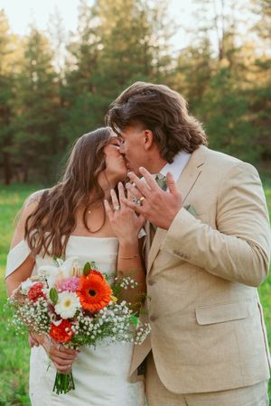 A romantic kiss shared between newlyweds holding a colorful bouquet in a forest setting.
