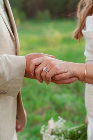 Close-up of newlyweds holding hands and showing engagement ring in natural outdoor setting.