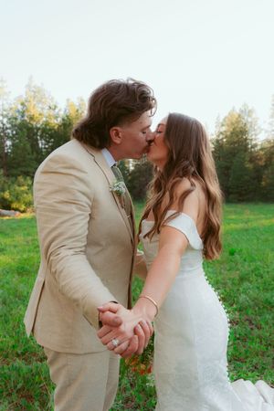 A tender wedding kiss shared in a picturesque outdoor setting with green grass and trees.