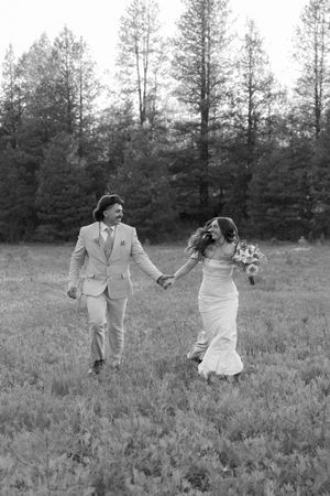 A couple walks hand in hand through a serene meadow with pine trees in the background, captured in black and white.
