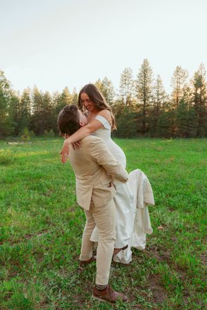 Couple performs playful lift pose during outdoor wedding photoshoot in grassy field.