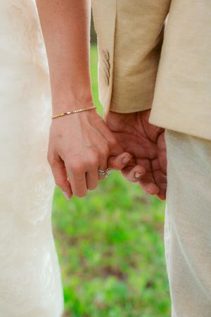 Two hands clasped together with a delicate bracelet and engagement ring against a soft green background.