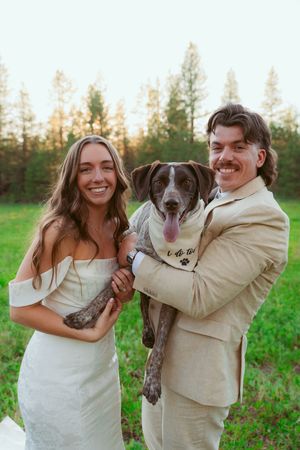 A joyful wedding portrait featuring the couple and their beloved dog in a sunny outdoor setting.