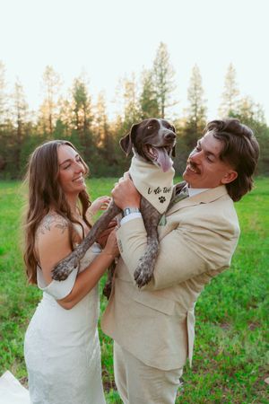 A happy couple in wedding attire holds their spotted dog in a grassy field with trees in the background at sunset.