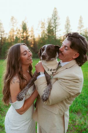 Newlyweds share playful kisses and laughs in a sunlit meadow wearing white dress and tan suit.