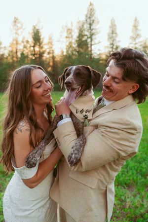 A joyful couple in wedding attire plays with their spotted dog in a grassy field during golden hour.