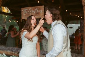 Couple shares a playful moment feeding each other wedding cake at rustic reception with twinkling lights and wooden sign.