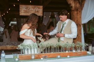 Newlyweds serve drinks together behind a bar decorated with white flowers during their wedding reception.