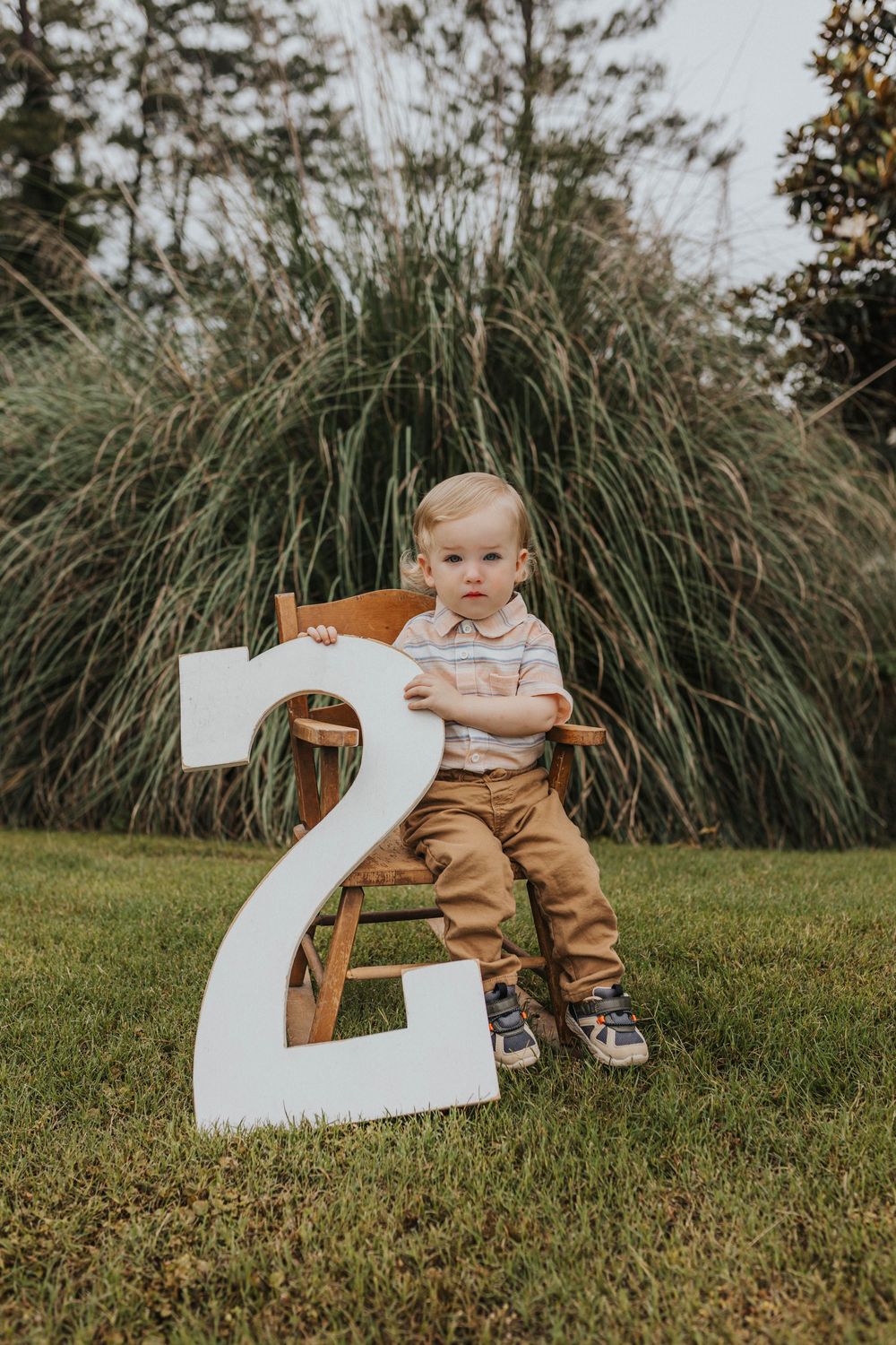 Toddler posing with large white number two on grass with ornamental grasses in background.